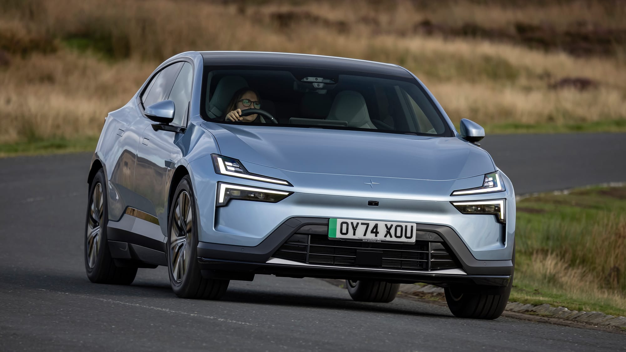 Imagem frontal de um Polestar 4 em tom azul claro, um SUV coupé elétrico projetado para alto desempenho e sustentabilidade. O design futurista é destacado pelos faróis em LED e a carroceria aerodinâmica. O cenário ao fundo é rural, com vegetação baixa e estrada pavimentada, enfatizando a versatilidade do veículo para diferentes ambientes. O Polestar 4 combina elegância, tecnologia e responsabilidade ambiental, refletindo o conceito de luxo sustentável.