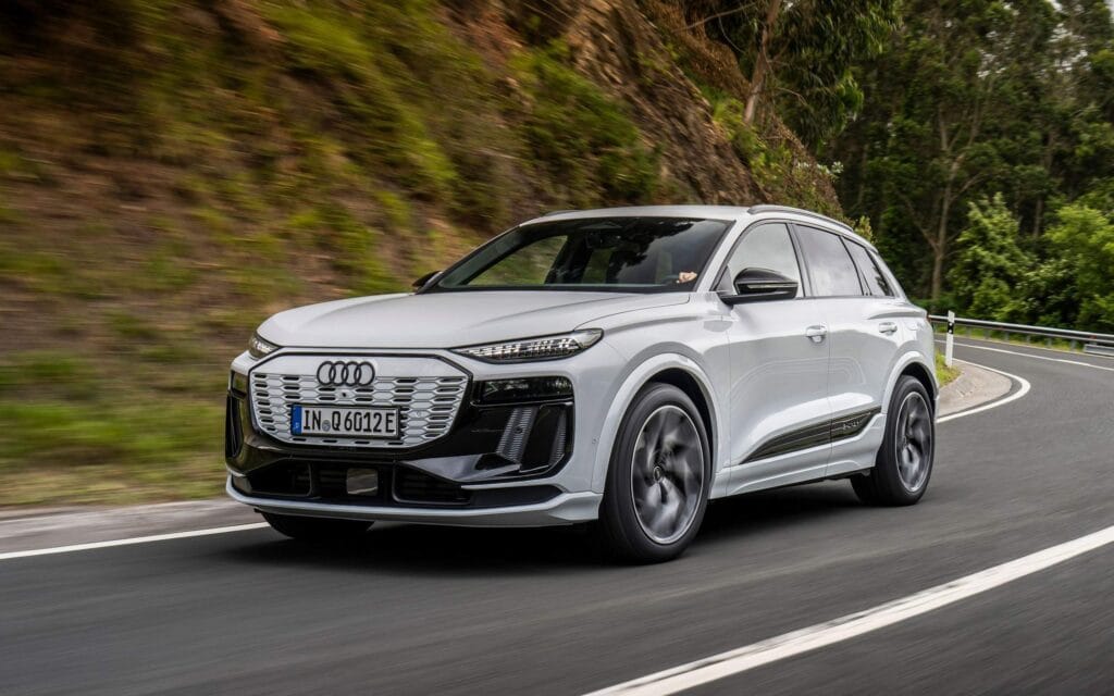 Imagem do Audi Q6 e-tron 2025 em movimento em uma estrada sinuosa, com vegetação densa ao fundo. Este SUV elétrico, na cor branca com detalhes em preto, exibe o design imponente e tecnológico característico da Audi, com uma grade frontal larga e faróis finos de LED. O Q6 e-tron combina desempenho e sustentabilidade, sendo ideal para consumidores que buscam um veículo de luxo com alta eficiência energética e conforto para viagens longas.